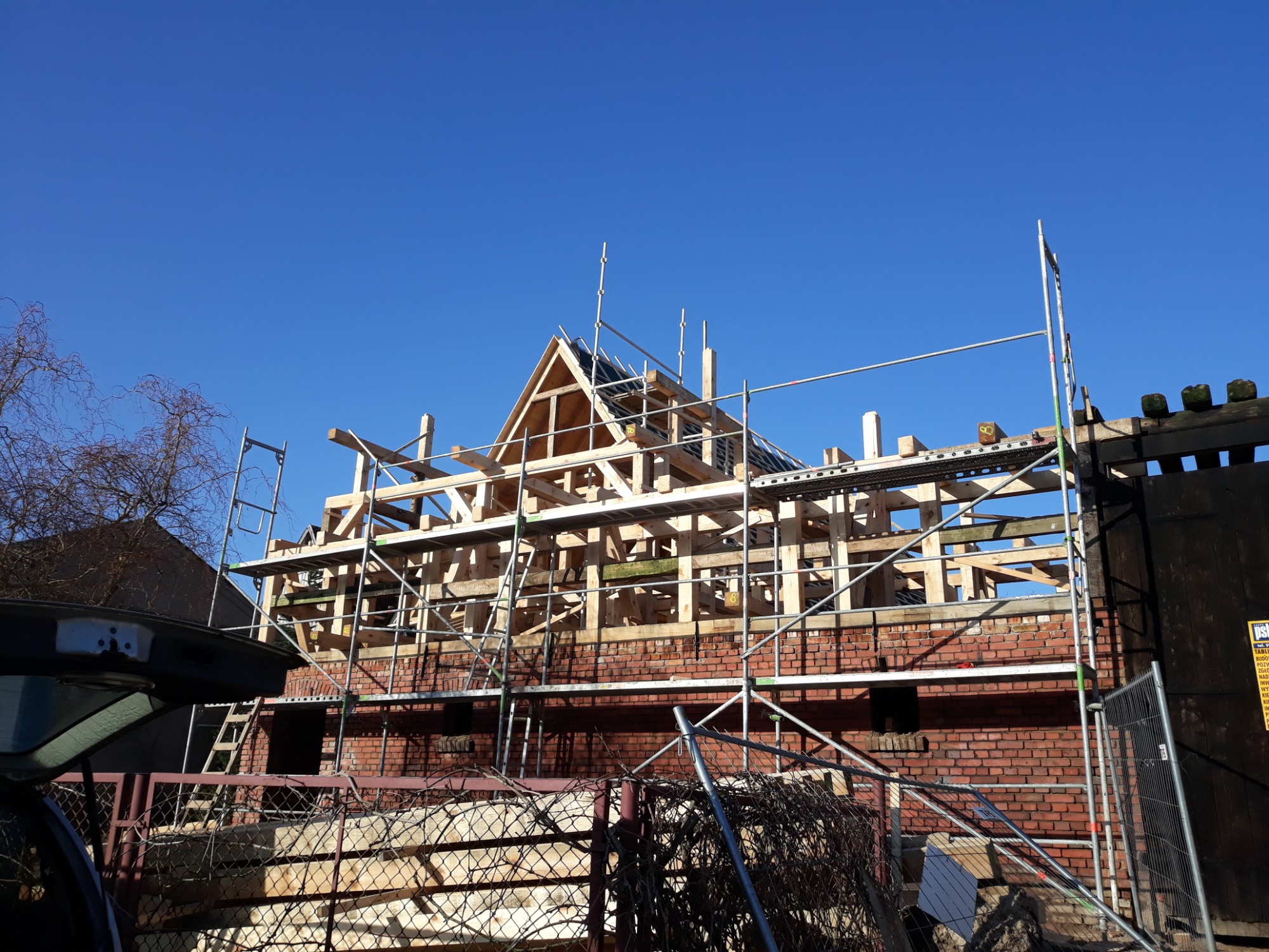 Budowa domu z czerwonej cegły w trakcie wznoszenia drewnianej konstrukcji dachu, widoczne rusztowania i błękitne niebo.