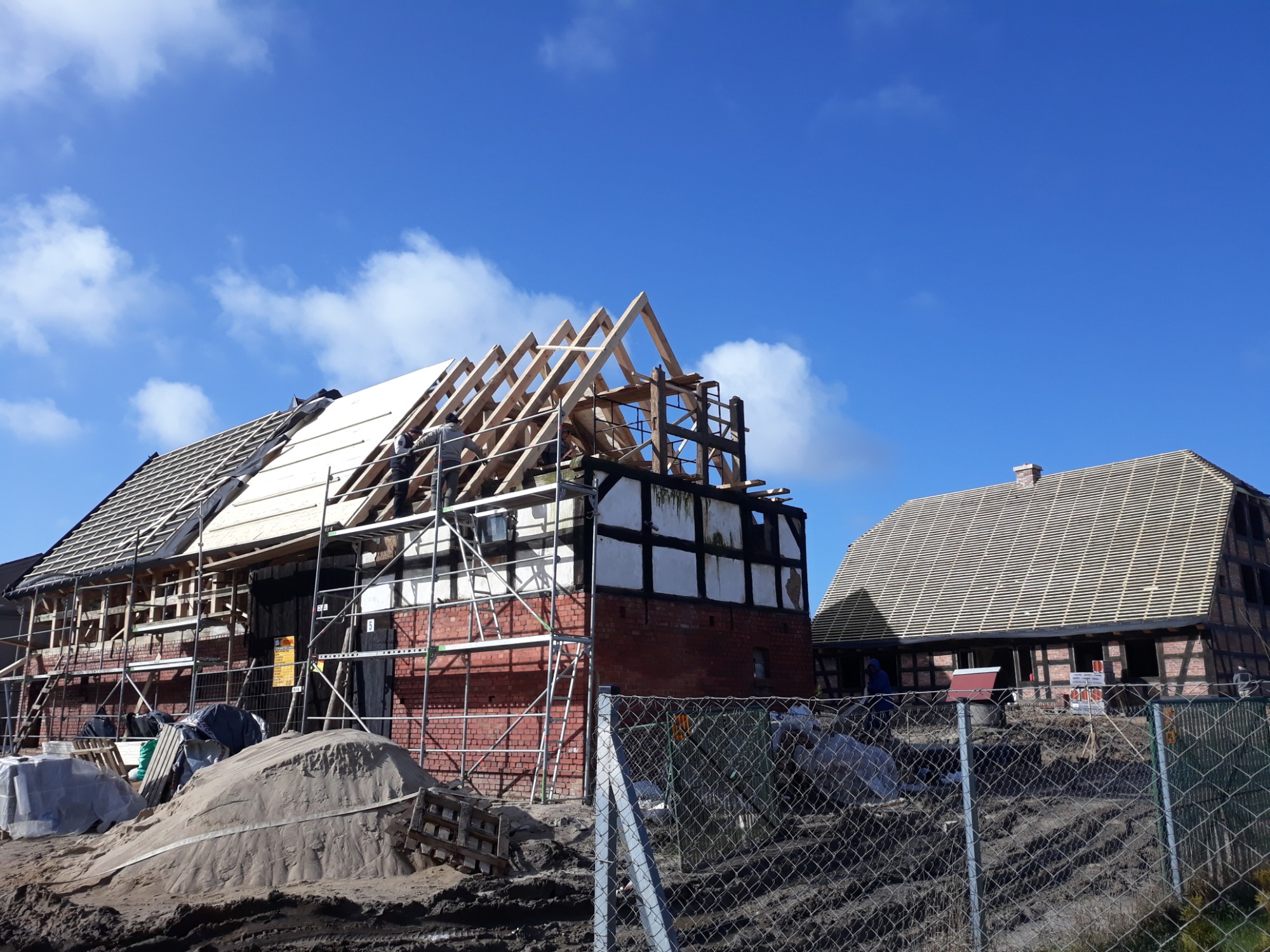 Budynek szkieletowy w trakcie renowacji dachu, widoczne rusztowania, drewniana konstrukcja więźby, częściowo pokryty dach deskami, w tle drugi budynek z nowym poszyciem dachowym, błękitne niebo...