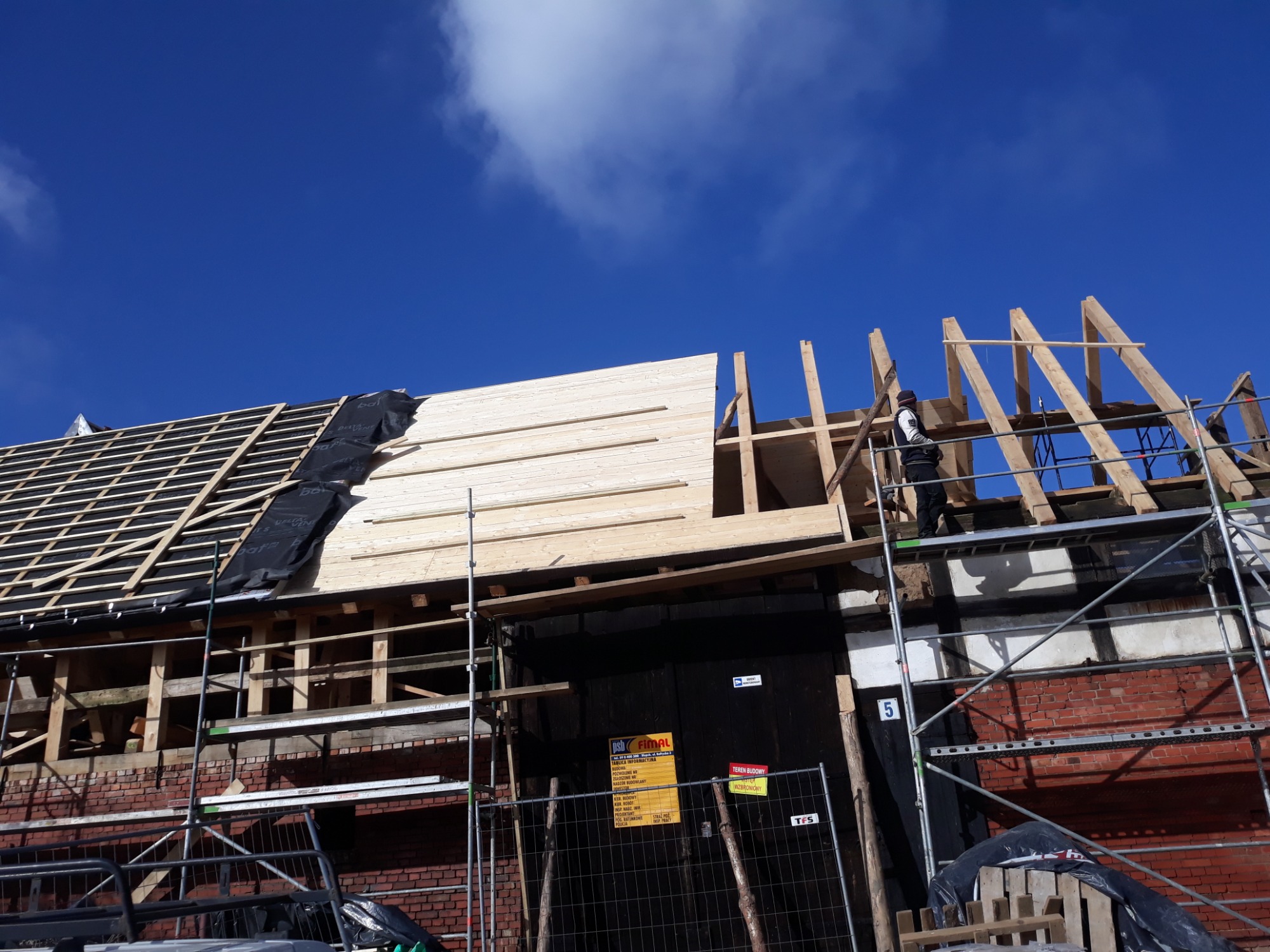 Montaż drewnianej konstrukcji dachu z widoczną więźbą i deskowaniem, częściowo pokrytą czarną folią, na tle błękitnego nieba z chmurami, z rusztowaniem i pracownikiem na górze.