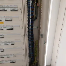 Otwarta szafa rozdzielcza z widocznymi bezpiecznikami, przewodami i listwami zaciskowymi, z przewodem uziemiającym po prawej stronie, zamontowana w jasnym pomieszczeniu.