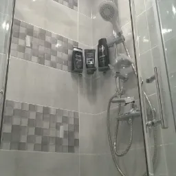 Szara kabina prysznicowa z deszczownicą, słuchawką prysznicową i półką na kosmetyki, wykończona płytkami w odcieniach szarości z dekoracyjnym pasem z motywem kostki.