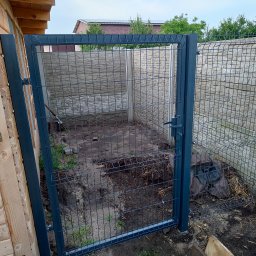 DM PARTNER Dariusz Pietrzak - Otwarta, grafitowa furtka z siatki ogrodzeniowej, przylegająca do drewnianej konstrukcji, z widokiem na betonowe elementy ogrodzenia i ziemię wewnątrz.