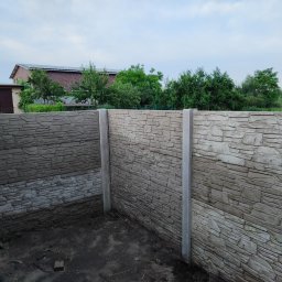 DM PARTNER Dariusz Pietrzak - Fragment nowo postawionego ogrodzenia z paneli imitujących kamień, widoczna szpadel oparta o słupek, w tle budynek z panelami solarnymi na dachu.