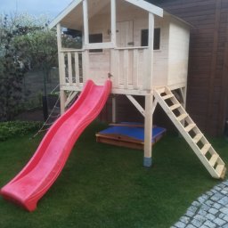 DM PARTNER Dariusz Pietrzak - Drewniany domek dla dzieci na podwyższeniu z czerwoną zjeżdżalnią i piaskownicą na trawniku.