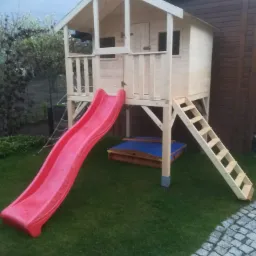 Drewniany domek dla dzieci na podwyższeniu z czerwoną zjeżdżalnią i piaskownicą na trawniku.
