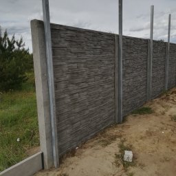 DM PARTNER Dariusz Pietrzak - Montaż prefabrykowanego ogrodzenia betonowego z metalowymi słupkami w trakcie instalacji, widoczna osoba pracująca przy dalszej części ogrodzenia.