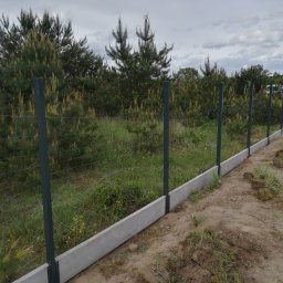 DM PARTNER Dariusz Pietrzak - Fragment nowo postawionego ogrodzenia z siatki, wspartego na betonowej podmurówce i metalowych słupkach, z widocznymi wykopanymi kępami trawy wzdłuż linii ogrodzenia i młodymi sosnami w tle.