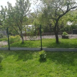 DM PARTNER Dariusz Pietrzak - Zielona siatka ogrodzeniowa otaczająca trawnik z młodymi drzewkami owocowymi i krzewami w słoneczny dzień.