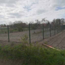 DM PARTNER Dariusz Pietrzak - Długa linia zielonej siatki ogrodzeniowej rozciąga się wzdłuż pola, z widocznymi słupkami i ziemią przygotowaną pod uprawę, na tle pochmurnego nieba i zabudowań.