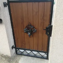 Brązowa furtka z drewnopodobnym wypełnieniem, czarną ramą i dekoracją z motywem winogron, zamontowana między dwoma słupkami.