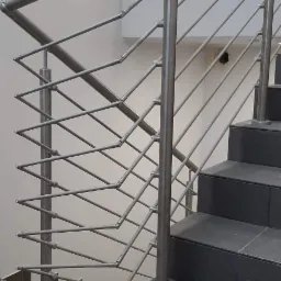 Schody z balustradą nierdzewną o geometrycznym wzorze wypełnienia, widoczne szare płytki podłogowe i fragment ściany.