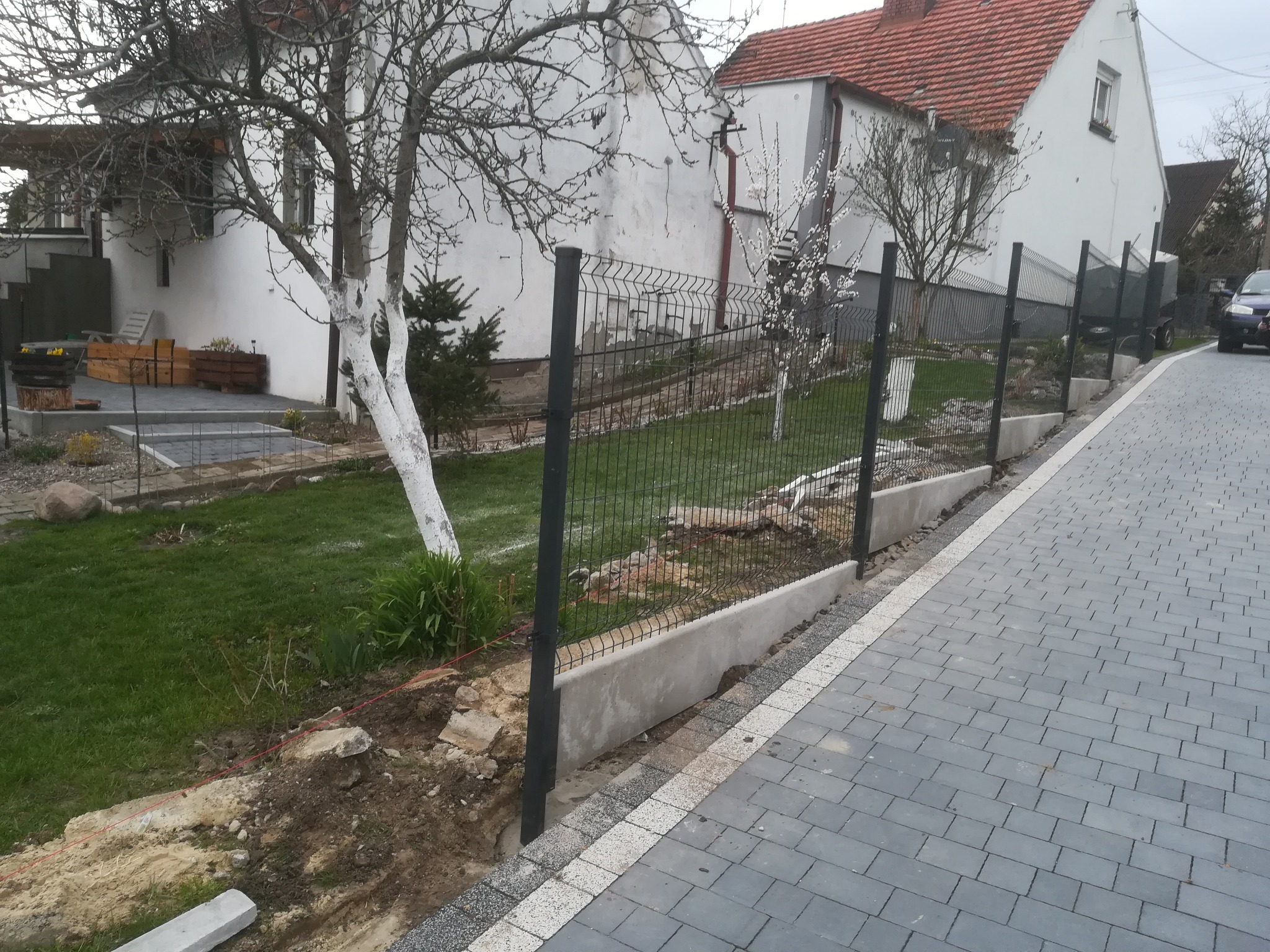 Etap budowy ogrodzenia panelowego z podmurówką betonową, widok z boku na posesję z kostką brukową i domem w tle.