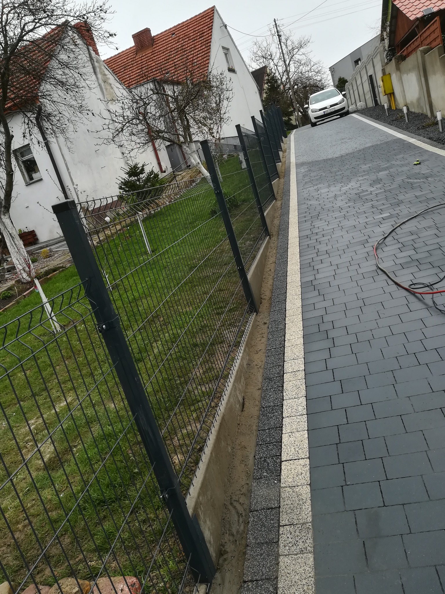 Nowoczesne, grafitowe ogrodzenie panelowe z widocznymi mocowaniami, oddzielające posesję od wybrukowanej drogi dojazdowej z kostki w odcieniach szarości i bieli, z domami w tle.