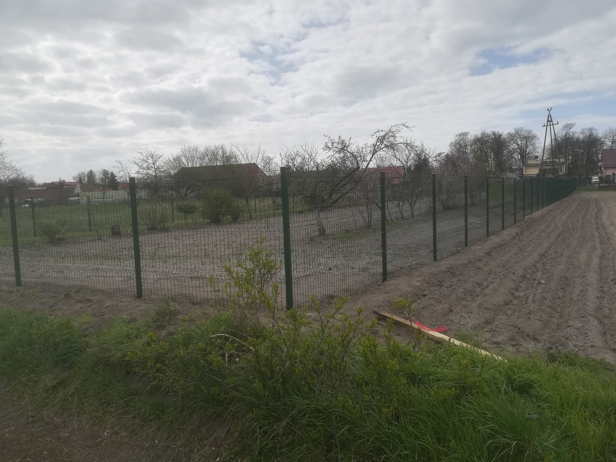 Długa linia zielonej siatki ogrodzeniowej rozciąga się wzdłuż pola, z widocznymi słupkami i ziemią przygotowaną pod uprawę, na tle pochmurnego nieba i zabudowań.