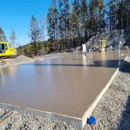 Świeżo wylana betonowa płyta w otoczeniu żwiru, z widocznym żółtym sprzętem budowlanym w tle i instalacją wodną na płycie, na tle lasu i błękitnego nieba.