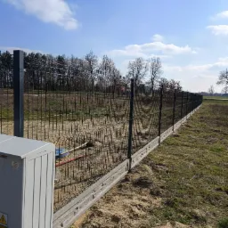 Długi, metalowy płot panelowy z podmurówką betonową, biegnący wzdłuż nierównego terenu z widocznymi śladami prac ziemnych, w tle drzewa i błękitne niebo z chmurami.