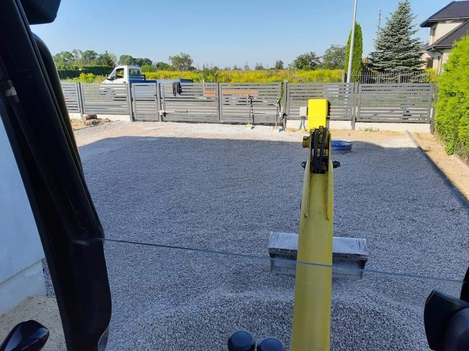 Widok z kabiny koparki na wyrównany teren posesji pokryty szarym żwirem; w tle nowoczesne ogrodzenie z metalowych paneli i zaparkowany biały bus.