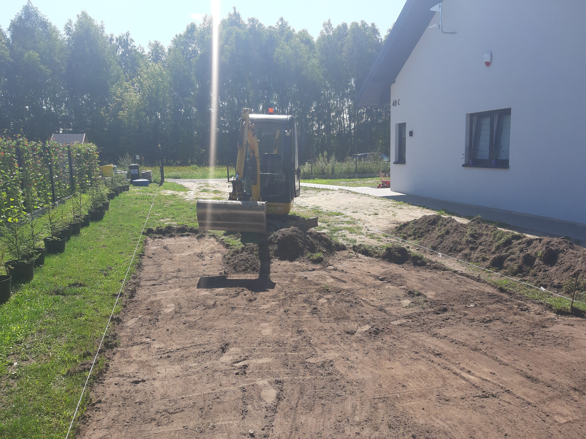 Żółta mini koparka na działce podczas prac ziemnych, widoczne wytyczone granice przyszłej instalacji, w tle budynek mieszkalny i drzewa.