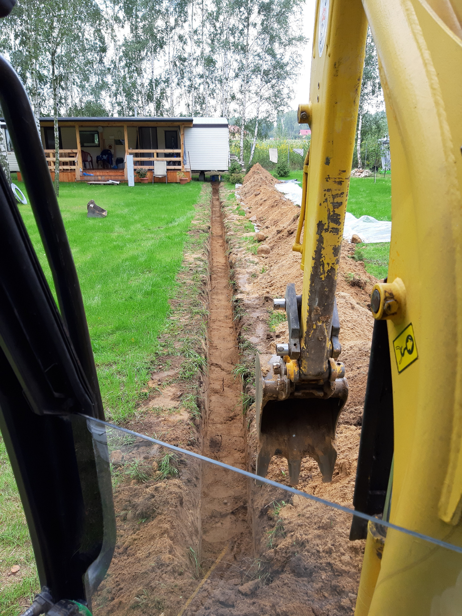 Wąski wykop liniowy w trawniku, widok z kabiny koparki, żółte ramię koparki częściowo zasłania widok, w tle domek letniskowy i drzewa.