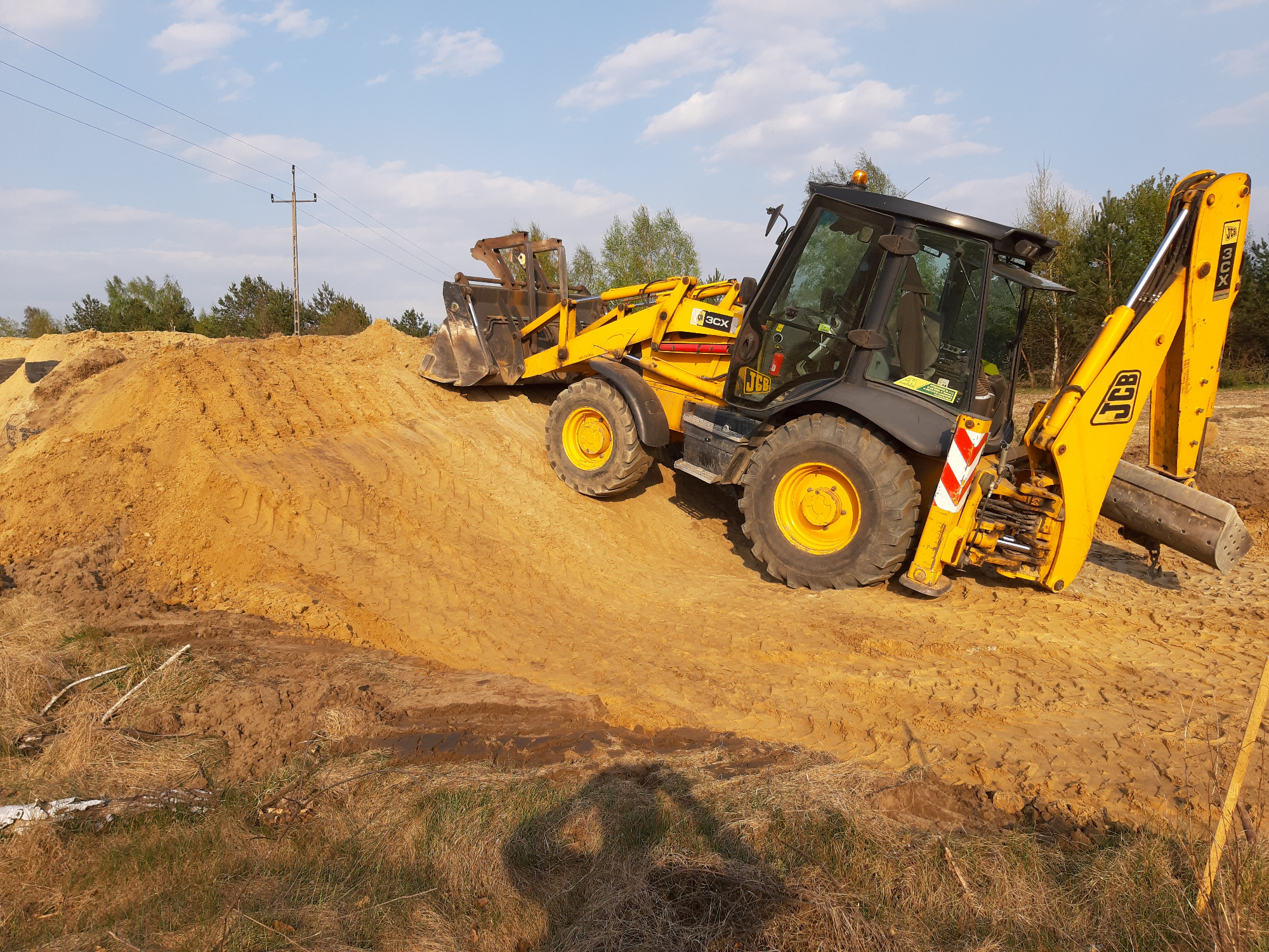 Żółto-czarna koparko-ładowarka JCB na nierównym terenie piaszczystym, z widocznym nasypem ziemi i elementami krajobrazu w tle.
