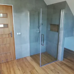 Nowoczesna kabina prysznicowa z odpływem liniowym, szare płytki na ścianach, drewnopodobna podłoga i drzwi z przeszkleniami w odnowionej łazience na poddaszu.