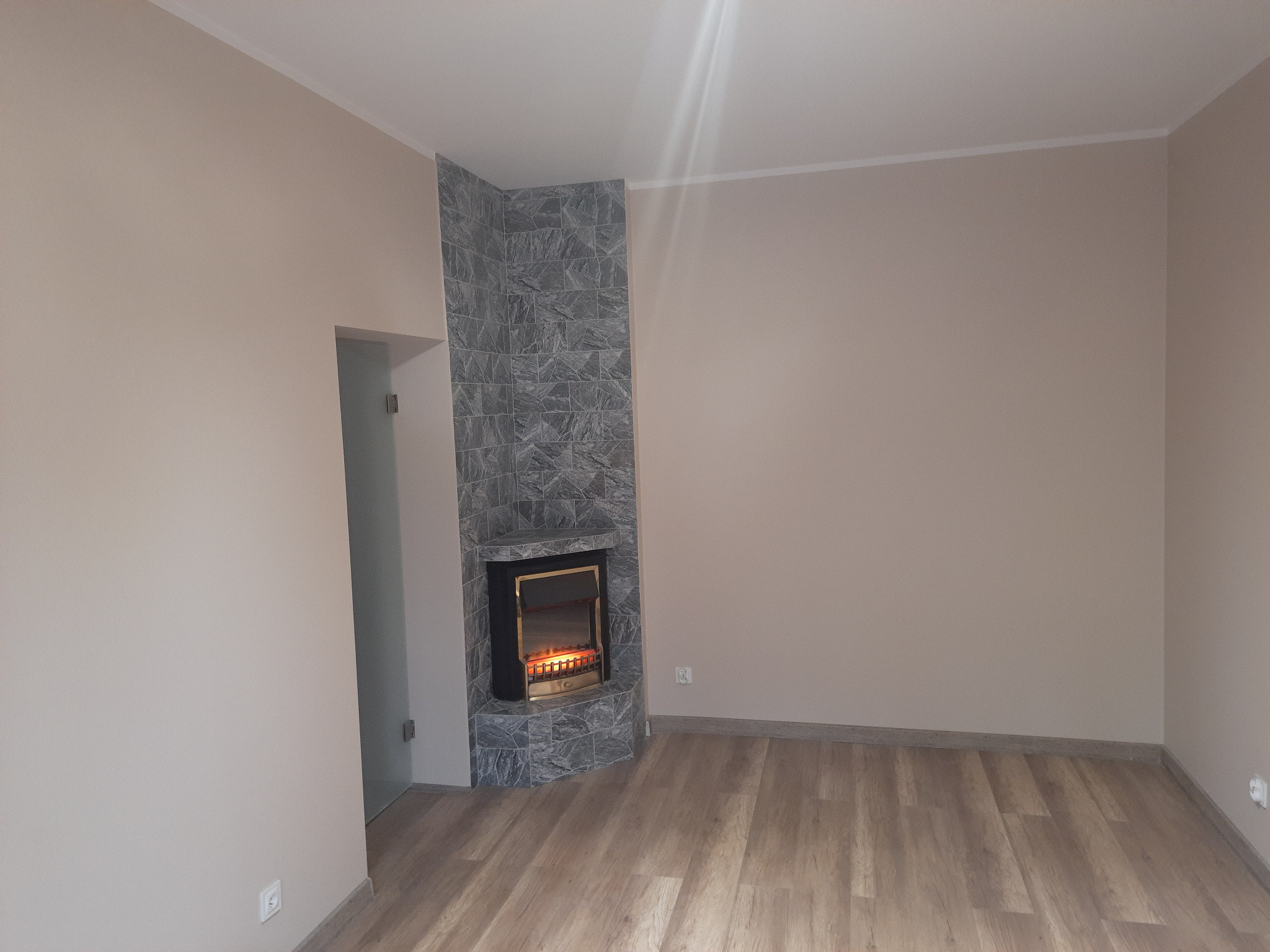 Salon po remoncie z kominkiem obłożonym szarą kamienną mozaiką, podłoga wyłożona panelami imitującymi drewno, ściany pomalowane na beżowo, widoczne gniazdko elektryczne.