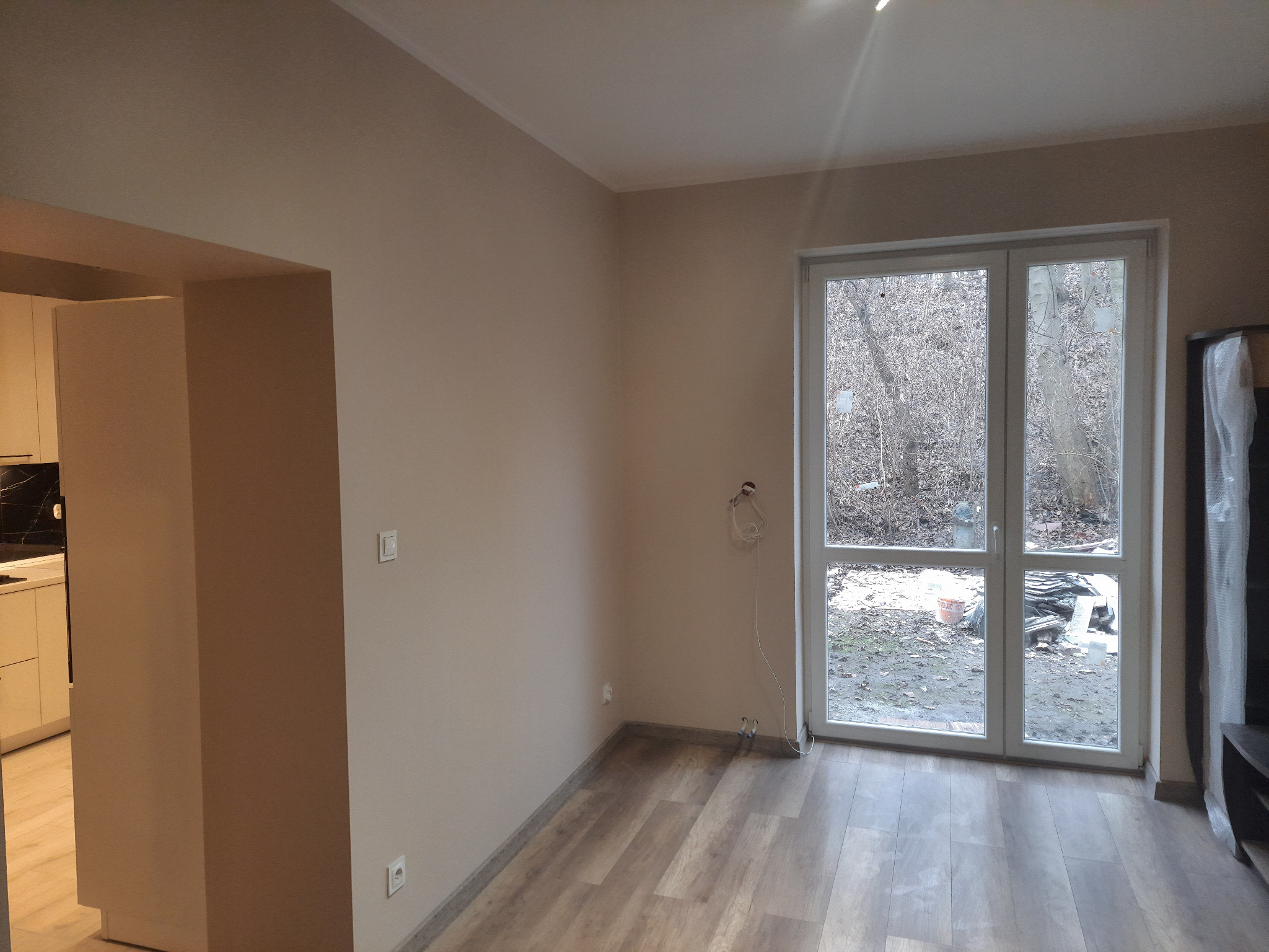 Wnętrze pokoju w Zabrzu po remoncie, z widoczną podłogą w panelach, świeżo pomalowanymi ścianami w odcieniach beżu, białymi drzwiami balkonowymi i fragmentem kuchni w tle.