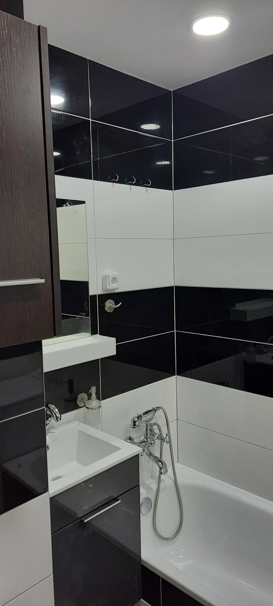 Nowoczesna łazienka z czarno-białymi płytkami ułożonymi w poziome pasy, białą umywalką w szafce z ciemnego drewna i chromowaną armaturą, widoczne haczyki na ścianie.