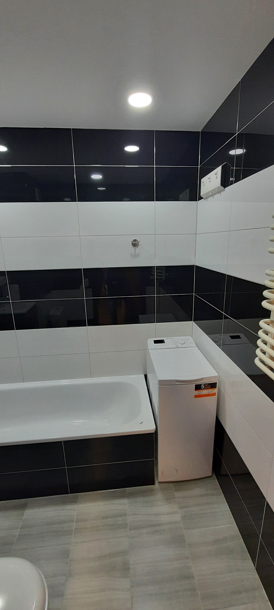 Nowoczesna łazienka z czarno-białymi płytkami ułożonymi w poziome pasy, białą wanną, pralką i grzejnikiem drabinkowym.