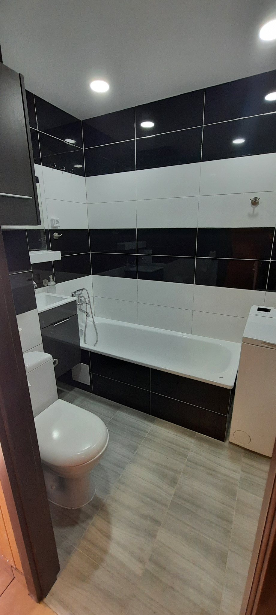 Nowoczesna łazienka z czarno-białymi płytkami ułożonymi w poziome pasy, białą wanną i toaletą oraz szarą szafką pod umywalką, oświetlona punktowymi lampami sufitowymi.