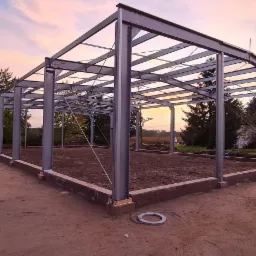 Konstrukcja stalowa hali w trakcie budowy, widoczne słupy i elementy szkieletu na tle zachodzącego słońca i drzew