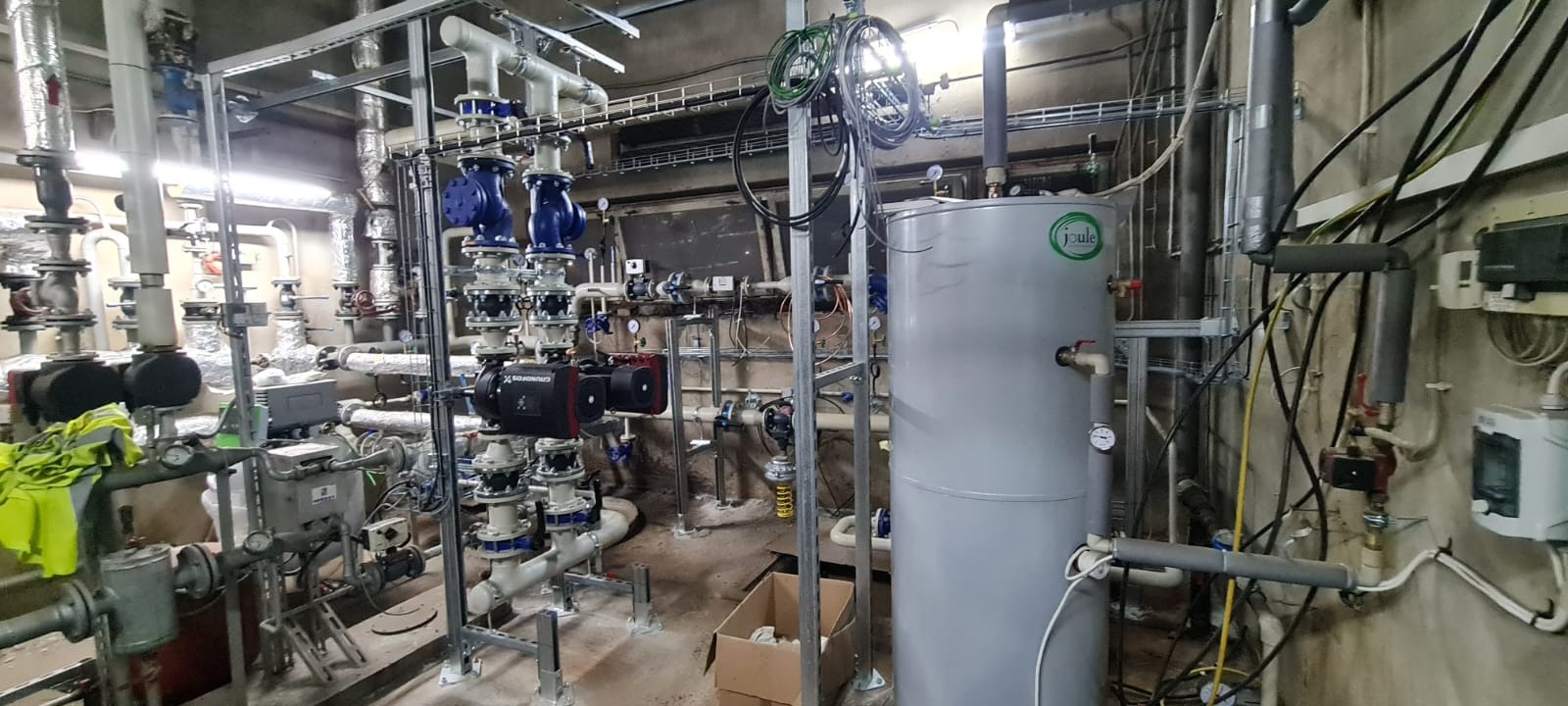 Złożona instalacja grzewcza z rurami, pompami Grundfos i zbiornikiem Joule w pomieszczeniu technicznym.