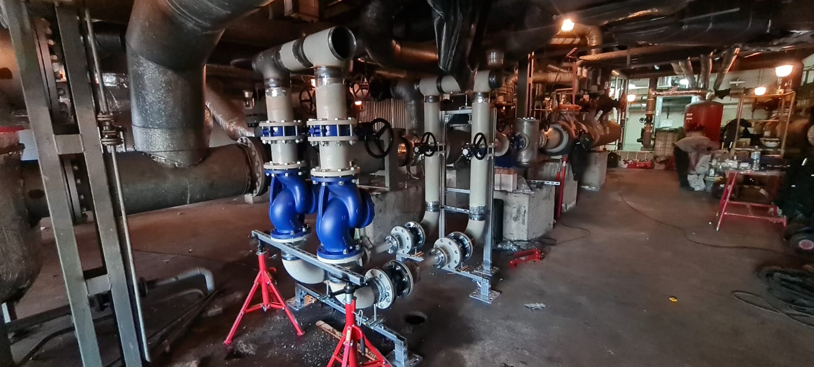 Instalacja przemysłowa z rurami, zaworami i niebieskimi pompami na czerwonych stojakach, widoczne spawane połączenia i osoba pracująca w tle.