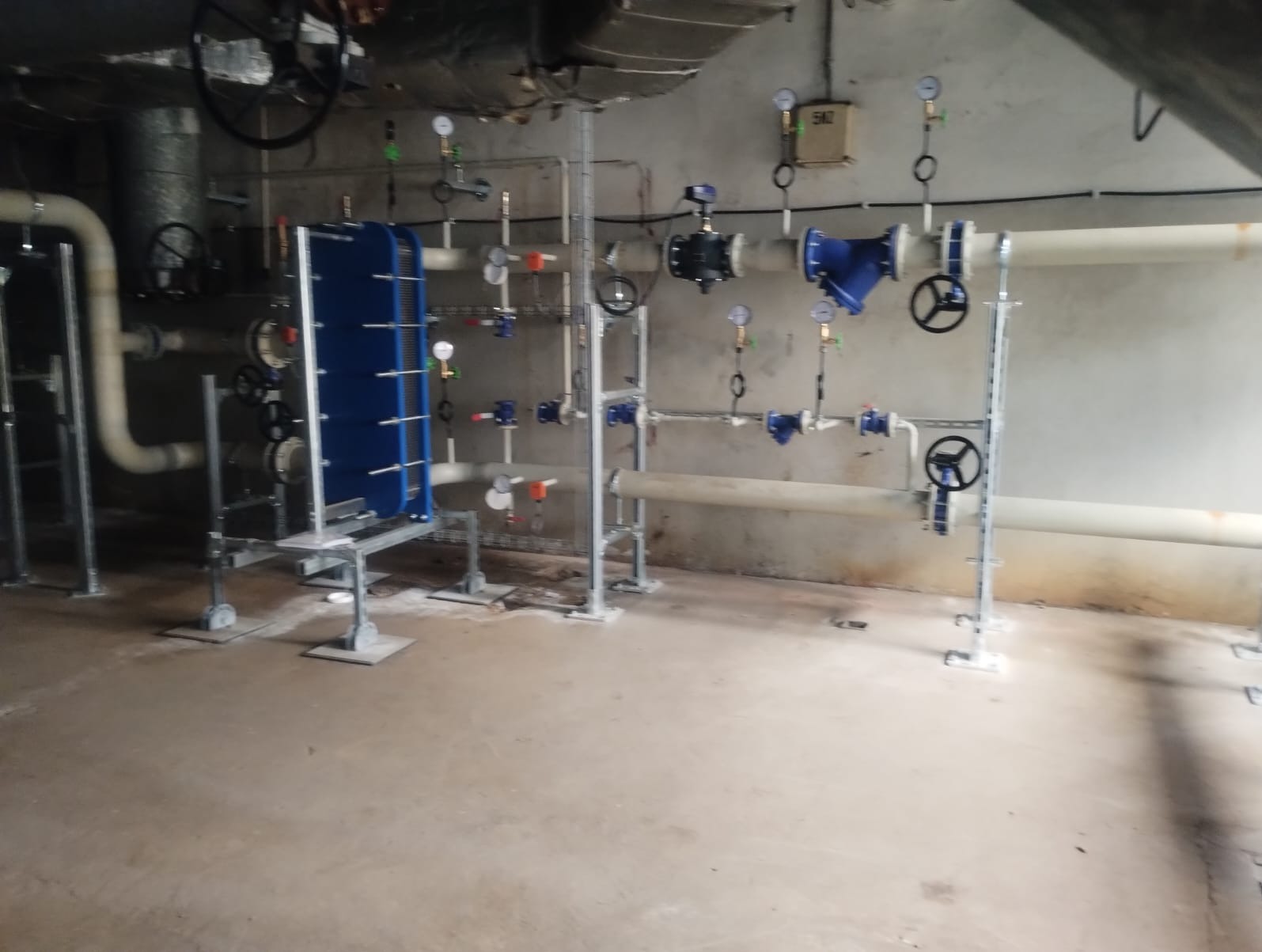 Instalacja przemysłowa z rurami, zaworami i wymiennikiem ciepła w kolorze niebieskim, zamontowana w hali z betonową podłogą i widocznymi elementami konstrukcyjnymi pod sufitem.