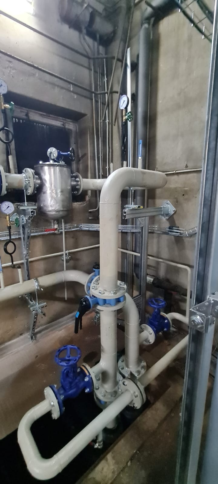 Instalacja przemysłowa z rurami, zaworami i manometrami w szarej, betonowej przestrzeni technicznej.