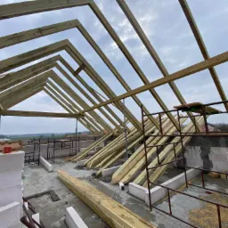 Wiązary dachowe z drewna na murowanym budynku w trakcie budowy, widok z dołu na konstrukcję dachu i pochmurne niebo.