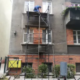 Pracownik w niebieskim kombinezonie na rusztowaniu czyści elewację budynku z widocznymi śladami po kulach, używając sprzętu do obróbki strumieniowo-ściernej; w tle ceglana część budynku z kwiatami...