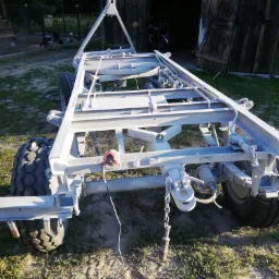 Rama przyczepy rolniczej po niedawnym pomalowaniu na jasny kolor, widoczne świeże ślady farby i zabezpieczony kabel, tło stanowi otwarte wrota stodoły i fragment podwórka.