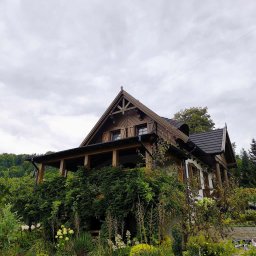 Usługi Stolarskie Oskar Pietras - Drewniany dom z zadaszonym tarasem, obrośniętym zielenią, w otoczeniu ogrodu. Widok z dołu, pochmurne niebo w tle. Charakterystyczna architektura.