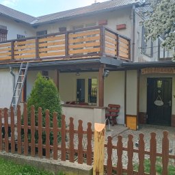Usługi Stolarskie Oskar Pietras - Dwukondygnacyjny dom z drewnianą balustradą na balkonie i ogrodzeniem z palików. Drabina oparta o balkon. Widoczny fragment kwitnącego drzewa.