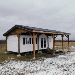 Usługi Stolarskie Oskar Pietras - Biały dom szkieletowy z drewnianą konstrukcją dachu na betonowych podstawach, otoczony polem z resztkami śniegu pod pochmurnym niebem.