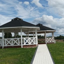 Usługi Stolarskie Oskar Pietras - Dwie białe altany z czarnym dachem, połączone ścieżką, na tle zielonego trawnika i pochmurnego nieba. Widoczny budynek z drewnianą elewacją w oddali.