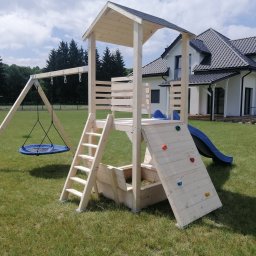 Usługi Stolarskie Oskar Pietras - Drewniany plac zabaw z huśtawką, zjeżdżalnią i ścianką wspinaczkową na trawniku przed domem. Jasne drewno, błękitne elementy. Widok z bliska, słoneczny dzień.