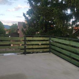 Usługi Stolarskie Oskar Pietras - Nowoczesna, zielona balustrada drewniana z metalowymi słupkami na betonowym tarasie. W tle zieleń drzew i zabudowa mieszkalna. Ujęcie z góry.
