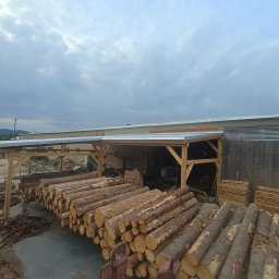 Usługi Stolarskie Oskar Pietras - Składowane bale drewna pod zadaszeniem z drewnianą konstrukcją. W tle budynek i pochmurne niebo. Widoczne deski i elementy konstrukcyjne.