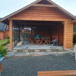 Usługi Stolarskie Oskar Pietras - Drewniana altana w trakcie budowy, widoczne narzędzia i materiały. Częściowo przeszklona ściana, surowe wykończenie podłogi. W tle dom z panelami słonecznymi.