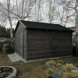 Drewniana, ciemnobrązowa altana z czarnym dachem bitumicznym, widoczna na tle drzew z białymi pniami i fragmentem żurawia budowlanego w tle, z paleniskiem z kamieni w pierwszym planie.