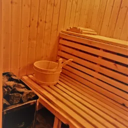 Wnętrze sauny z drewnianym wykończeniem, ławkami, wiadrem i piecem z kamieniami.