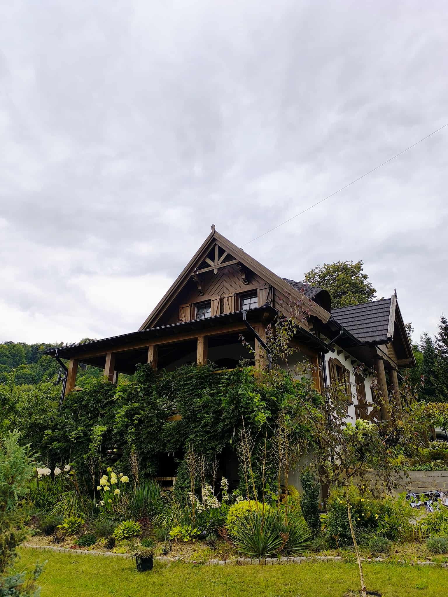 Drewniany dom z zadaszonym tarasem, obrośniętym zielenią, w otoczeniu ogrodu. Widok z dołu, pochmurne niebo w tle. Charakterystyczna architektura.