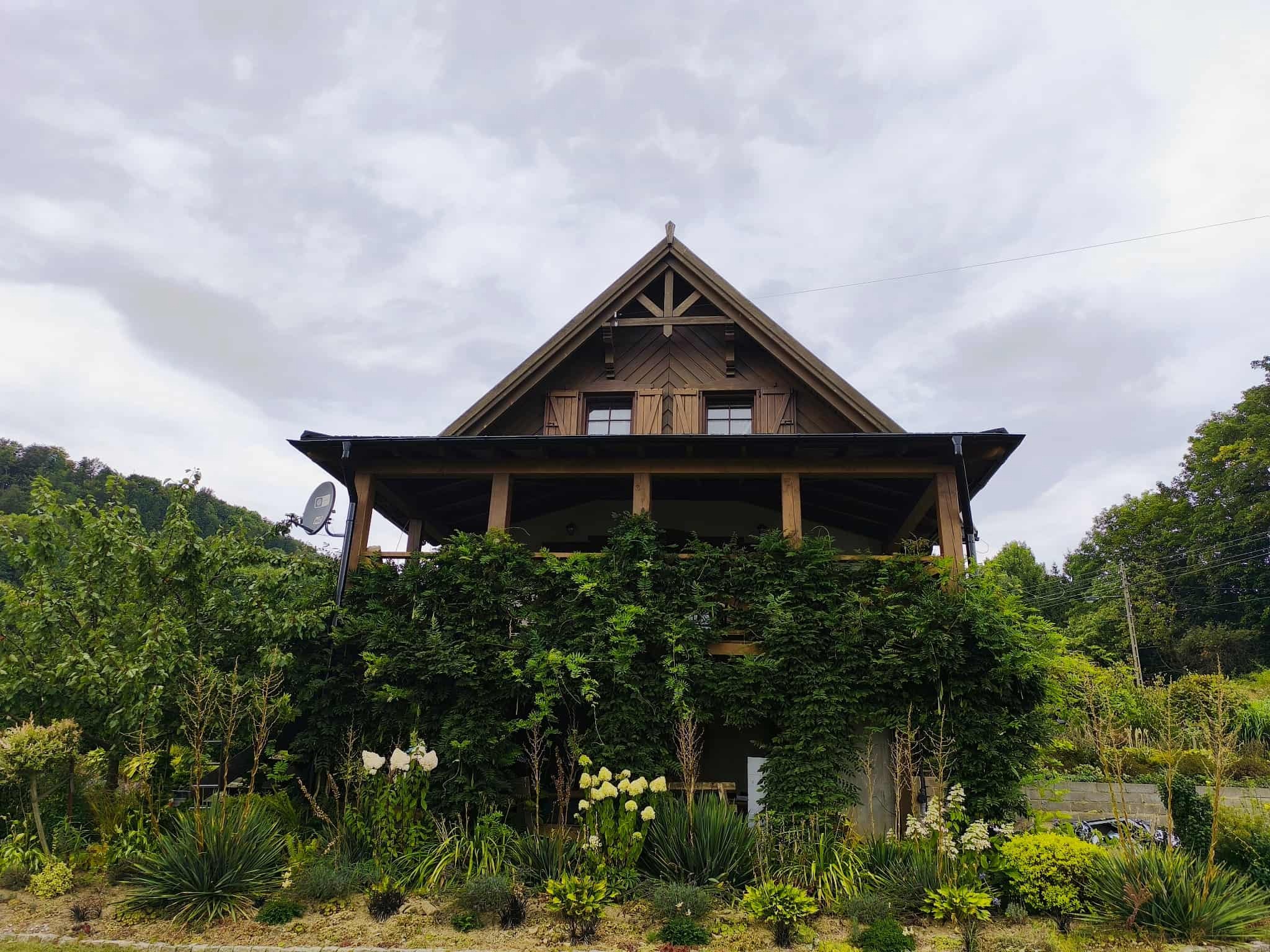 Drewniany dom z poddaszem i zadaszonym tarasem, porośniętym zielenią. Widok z dołu na budynek z dekoracyjnym szczytem i drewnianymi okiennicami. W tle pochmurne niebo.